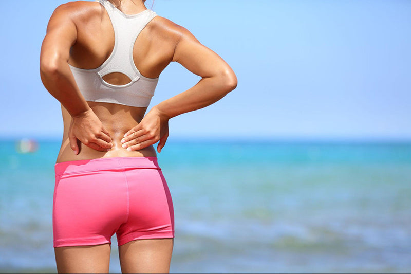 A woman in workout clothing faces the ocean and presses both hands to her lower back.