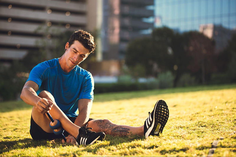 A man wearing workout clothing sitting in the grass using a MOBO tool to perform myofascial release on his right calf.