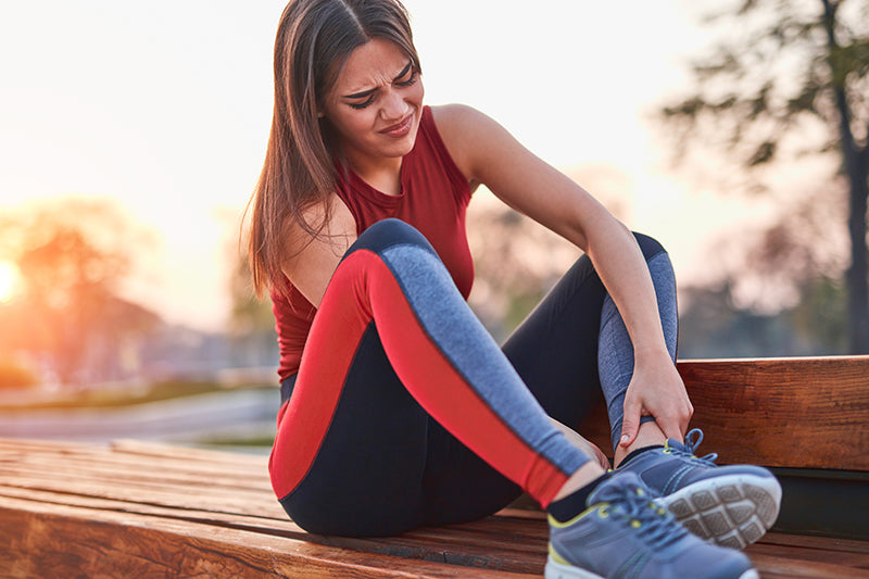 A woman in workout clothing sits and rubs her sore ankle.